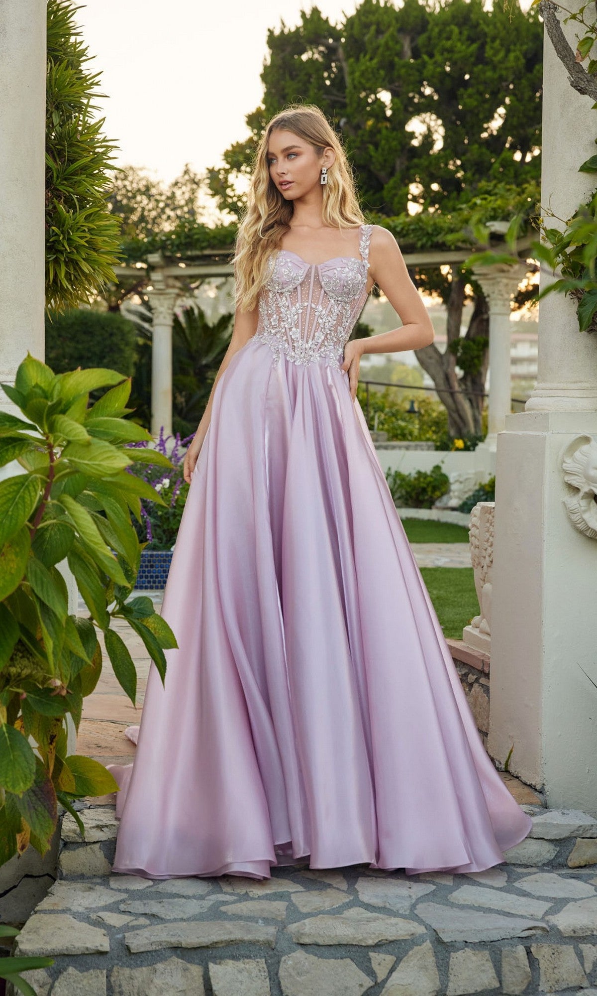 Amid lush greenery and stone columns, a woman stands outdoors in the Juliet JT2639G—a light purple satin a-line prom dress with a corset bodice, beaded applique, and cap sleeves.